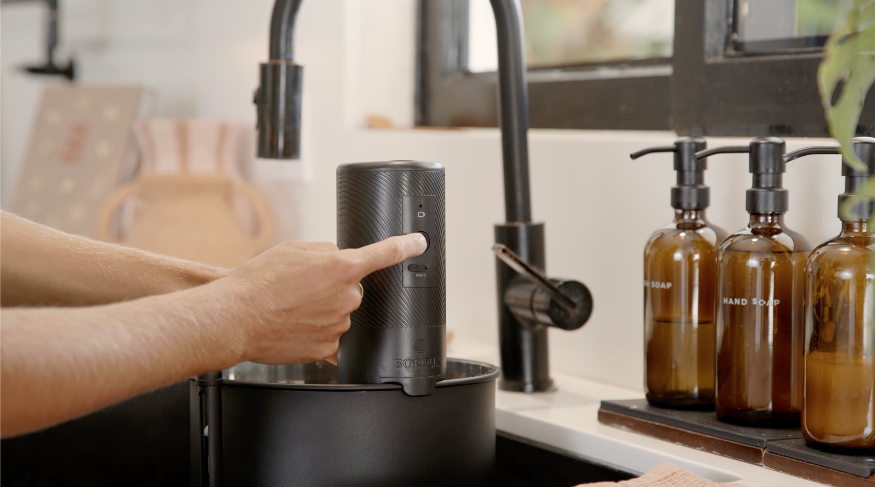 Person using a black kitchen appliance near a sink with brown bottles in the background